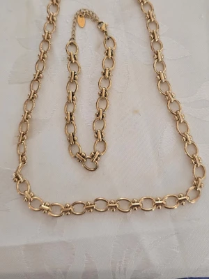 Guldfärgat kedjehalsband rostfritt stål  - Snyggt halsband i guldfärgad metall med stora, ovala länkar och dekorativa detaljer mellan varje länk. Kedjan har ett klassiskt och trendigt utseende som passar till många olika stilar. Justerbar längd med karbinlås och förlängningskedja.