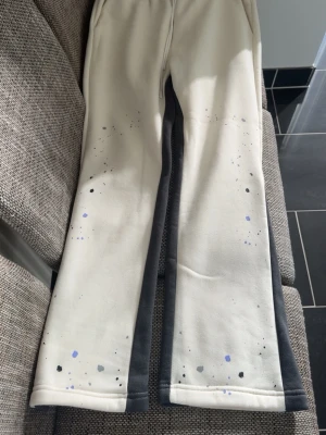 Beige mjukisbyxor med färgstänk - Säljer ett par beige mjukisbyxor med svarta sidopaneler och coola färgstänk i svart och lila. Byxorna har resår i midjan och raka ben. Perfekta för en avslappnad och trendig look.