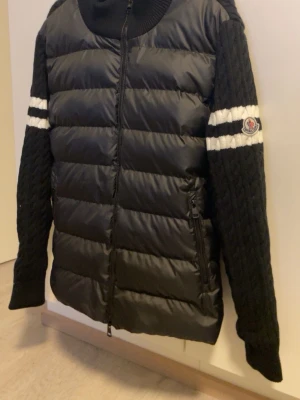  Moncler Cardigan - Snygg svart pufferjacka från Moncler med stickade ärmar och vita ränder på armarna. Jackan har dragkedja framtill, två fickor med dragkedja och Moncler-logga på ena ärmen. Kroppen är quiltad och vadderad medan ärmarna är kabelstickade.