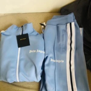 Palm Angels ljusblå mjukisbyxor - Snygga ljusblå mjukisbyxor från Palm Angels med vita och svarta ränder längs sidorna. Byxorna har resår i midjan och sidofickor, samt Palm Angels-logga tryckt på benet. Perfekta för en chill och sportig look.