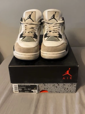 Air Jordan 4 Retro vit/grå/svart - Säljer ett par Air Jordan 4 Retro sneakers i vitt, svart och neutralgrått. Klassisk siluett med meshpaneler, mockadetaljer och ikonisk Jumpman-logga på plösen. Skorna har en chunky sula och coola detaljer som gör dem till ett måste för sneakerheads.