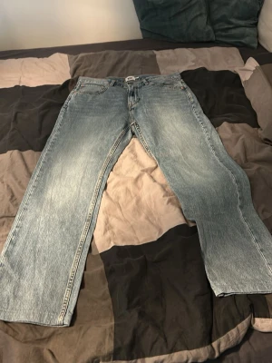 Ljusblå raka jeans från 157 - Säljer ett par ljusblå jeans från 157 med rak passform och klassisk femficksdesign. Jeansen har slitna detaljer och kopparfärgade nitar. Tillverkade i denim med bälteshällor och knappgylf. Perfekta för en avslappnad och trendig look.