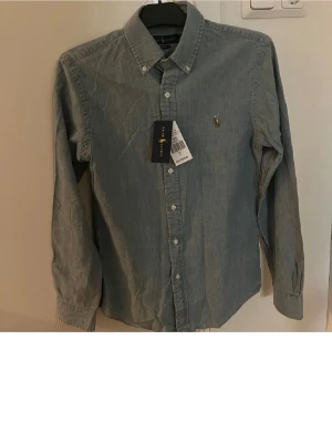 Blå skjorta från Ralph Lauren - Klassisk blå skjorta från Ralph Lauren med button-down krage och broderad logga på bröstet. Skjortan är långärmad och har knappar framtill. Perfekt för en stilren look och tillverkad i mjuk bomull.