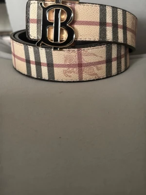 Beige rutigt  bälte - Snyggt bälte med klassiskt beige, svart och rött rutmönster. Bältet har ett stort guldfärgat B-spänne som ger en lyxig känsla. Perfekt accessoar för att lyfta din outfit och visa din stil.