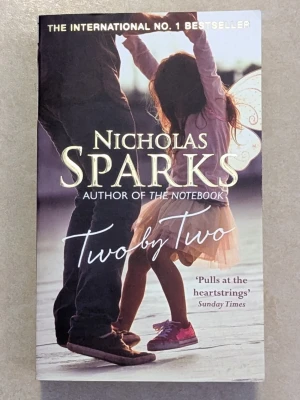Two by Two av Nicholas Sparks - En internationell bästsäljare av Nicholas Sparks, författaren till The Notebook. Omslaget visar en vuxen och ett barn som dansar tillsammans, vilket speglar bokens känslosamma och relationella tema. Pocketbok med mjukt omslag i färgerna rosa, lila och beige. Pocketboken är på engelska 