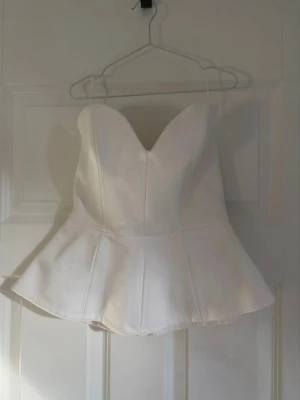 Vit peplum korsettopp - Säljer en vit korsettopp med peplumskärning och sweetheart-urringning. Toppen är axelbandslös och har en dragkedja i ryggen för enkel på- och avtagning. Perfekt för att skapa en snygg siluett och passar till många olika looks.