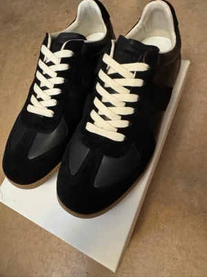  Maison Margiela Gats - Svarta sneakers från Maison Margiela med klassisk design, vita snören och beige gummisula. Skorna har en mix av mocka och läder på ovandelen samt vit insida. Perfekta för dig som gillar stilrena och tidlösa sneakers. Helt oanvända och nya 