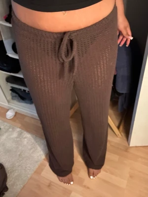 Bruna stickade byxor med knytband - Säljer ett par bruna stickade byxor med bred resår och knytband i midjan. De har en lös passform och är tillverkade i ett genombrutet, mjukt material som ger en chill och trendig vibe. Perfekt nu för sommaren, de är en L men passar M som ni ser på bilden. Jag har bara provat dom aldrig använt 