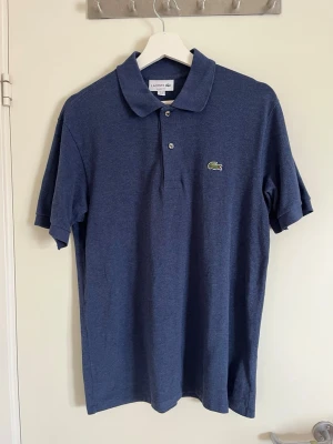 Oanvänd blå Lacosté piké - Klassisk blå pikétröja från Lacoste med korta ärmar och krage. Tröjan har knappar framtill och den ikoniska gröna krokodilloggan på bröstet. Perfekt för en stilren och avslappnad look.