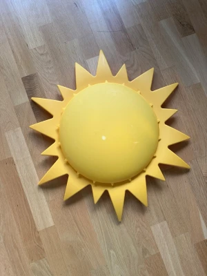Gul solformad taklampa IKEA - Säljer en solformad taklampa från IKEA i plast. Lampan är gul och har en rund mitt med taggiga solstrålar runt om. Perfekt som dekorativ belysning till barnrum eller som en rolig detalj i hemmet. Lampan har plats för två glödlampor.