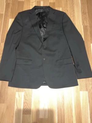 Svart kavaj Dressmann Slim Fit - Stilren svart kavaj från Dressmann i modellen Slim Fit. Kavajen har två knappar framtill, klassisk krage och fickor med lock. Fodret är svart med röda detaljer på insidan. Perfekt för en dressad look.