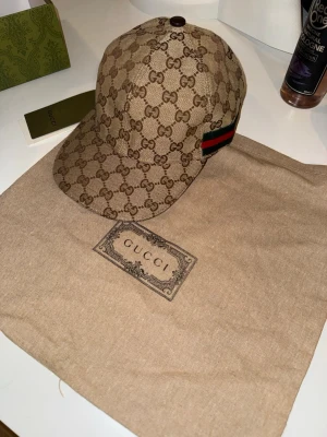 Beige keps från Gucci med GG-mönster - Säljer en beige keps från Gucci med det klassiska GG-monogrammönstret över hela kepsen. På sidan finns en grön och röd rand som signaturdetalj. Kepsen är tillverkad i canvasmaterial och har en böjd skärm. Perfekt för dig som vill ha en lyxig och trendig accessoar.
