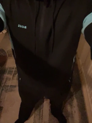 Svart och ljusblå BOSS loungewear set - Säljer ett svart loungewear set från BOSS med ljusblå detaljer på ärmar och sidor. Setet består av en hoodie med dragsko och matchande byxor. Perfekt för chill dagar hemma. Materialet känns mjukt och bekvämt mot huden.