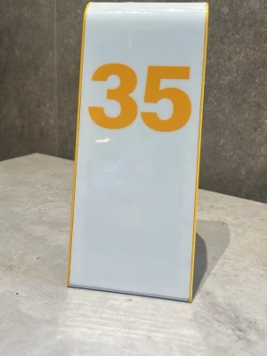 Bordsskylt med siffran 35 - Vit bordsskylt med stora gula siffror '35' och gula kanter. Skylten är rektangulär och står upp på bordet, perfekt för att markera nummer vid till exempel restaurangbord eller event.
