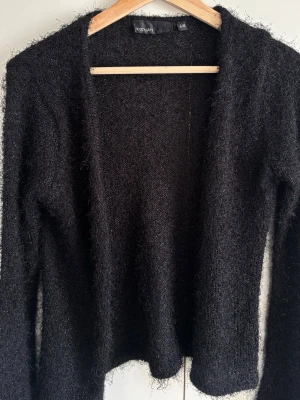 Svart kofta från KappAhl - Mysig svart kofta från KappAhl med fluffig textur och glitter aktit i tyget. Långärmad modell som är perfekt att slänga på sig över en t-shirt eller topp. Koftan har en enkel och stilren design som passar till många olika outfits.