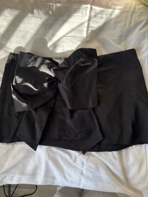 Svart kortkjol med stor rosett H&M - Säljer en svart kortkjol från H&M med en stor, iögonfallande rosett framtill. Kjolen har en elegant och stilren look. Strl 46 men passar ca M som en lågmidjad kjol