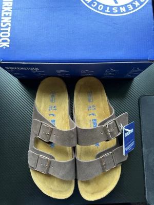 Birkenstock Arizona sandaler mocka brun - Klassiska Birkenstock Arizona sandaler i brun mocka med två breda remmar och justerbara metallspännen. Fotbädden är i kork och mocka för extra komfort. Sulan är räfflad och ger bra grepp. Perfekta för en avslappnad och trendig stil.