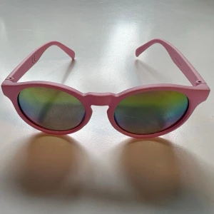 Runda rosa solglasögon - Säljer ett par riktigt snygga solglasögon med rund form och rosa bågar. Glasen har en spegelreflex som skiftar i grönt och blått. Perfekta för att sticka ut och ge din look en färgklick i sommar.