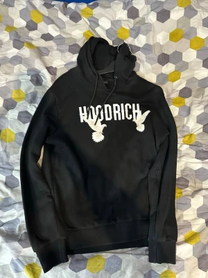 Svart hoodie från Hoodrich med tryck - Svart hoodie från Hoodrich med vit logga och två vita duvor tryckta på bröstet. Klassisk huva med dragsko och ribbade muddar. Perfekt för dig som gillar streetwear och vill ha en stilren men ändå unik look.
