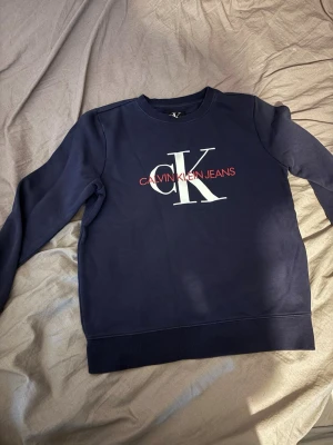 Mörkblå sweatshirt Calvin Klein Jeans - Mörkblå sweatshirt från Calvin Klein Jeans med stort vitt CK-tryck och röd text på bröstet. Klassisk rund halsringning och ribbade muddar vid ärmslut och nederkant. Perfekt för en avslappnad och stilren look.För barn i ca 12-års åldern