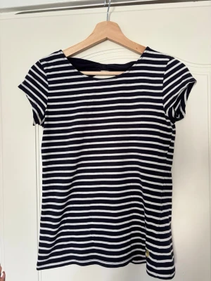 Randig t-shirt med rosett i ryggen - Snygg t-shirt med marinblå och vita ränder samt en söt rosettdetalj i ryggen. T-shirten har korta ärmar och rund halsringning. Perfekt för dig som gillar klassisk stil med en twist. Den har några defekter så som några få fläckar som knappt märks. Lösa trådar och svettfläckar 