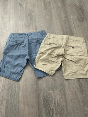 Två par shorts i blått och beige - Säljer två par klassiska shorts, ett par i ljusblått med diskret rutmönster och ett par i beige. Båda har knapp- och gylfstängning, sidofickor och bakfickor med knapp. Perfekta för varma dagar och enkel stil.