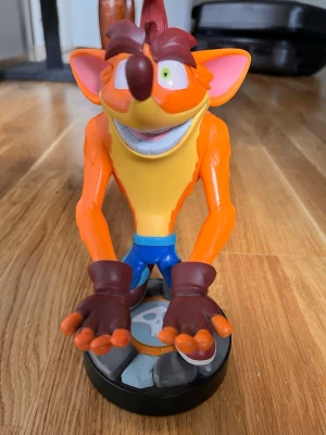 Crash Bandicoot spelkontrollerhållare - Cool hållare för spelkontroller i form av Crash Bandicoot. Figuren är färgglad med orange kropp, blå byxor och röda skor, står på en rund svart bas. Perfekt för att förvara din handkontroll eller mobil på ett snyggt sätt i gamingrummet.