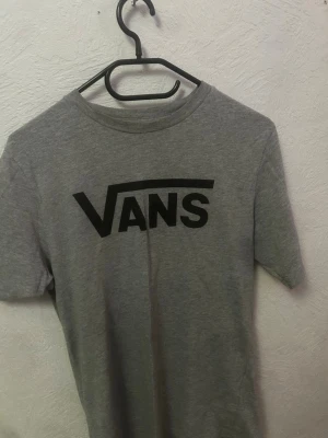 Grå Vans t-shirt med logga - Snygg grå t-shirt från Vans med den klassiska svarta loggan tryckt framtill. T-shirten har rund hals och korta ärmar, perfekt för en avslappnad streetwear-look. Tillverkad i mjuk bomull för skön känsla hela dagen.