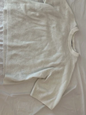 Vit stickad tröja från 157 - Mysig vit stickad tröja från 157 med korta ärmar och ribbade detaljer vid hals, ärmslut och nederkant. Tröjan har en avslappnad passform och är perfekt för lager på lager. Materialet känns mjukt och bekvämt mot huden.
