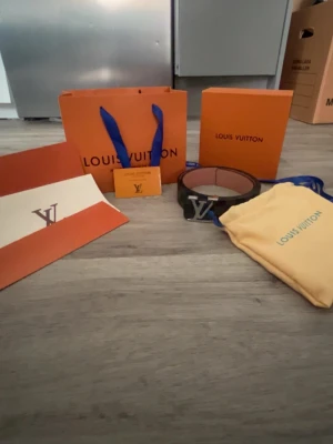 Louis Vuitton rutigt svart bälte - Snyggt svart bälte från Louis Vuitton med klassiskt rutigt mönster och stor LV-logga i silver på spännet. Bältet är gjort i skinn och har en exklusiv känsla, perfekt för att lyfta vilken outfit som helst.