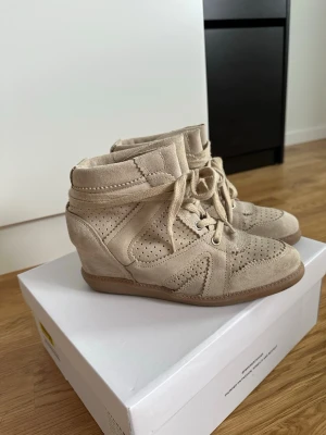 Beige sneakers i mocka - Snygga beige sneakers med kilklack och perforerade detaljer i mjuk mocka. Skorna har snörning framtill och en högre siluett som ger extra attityd till din outfit. Perfekta för dig som vill ha både stil och komfort i ett par sneakers.