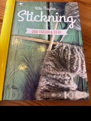 Stickning: 200 frågor & svar - En inspirerande bok om stickning av Rita Taylor. Omslaget har en grön träbakgrund med stickor och grått garn i fokus. Perfekt för dig som vill lära dig mer om stickning och få svar på vanliga frågor.