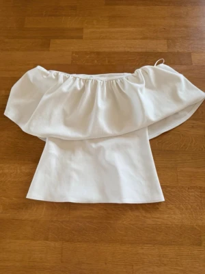 Vit offshoulder topp med volang - Säljer en snygg vit offshoulder topp med bred volang runt axlarna. Toppen är ärmlös och har en figurnära passform. Perfekt för dig som vill ha en stilren och trendig look.