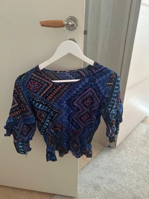 Blå mönstrad blus med volang - Säljer en blå blus med färgstarkt geometriskt mönster och volangdetaljer. Blusen har trekvartsärm med knyt vid ärmslut och en croppad passform. Perfekt för dig som gillar unika toppar med mycket färg och detaljer!💙💜
