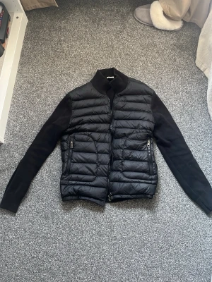 Moncler cardigan - Snygg svart cardiganjacka från Moncler med quiltad front och stickade ärmar. Jackan har dragkedja framtill, två fickor med dragkedja och en liten Moncler-logga på ena ärmen. Perfekt för dig som gillar stilren och modern design.