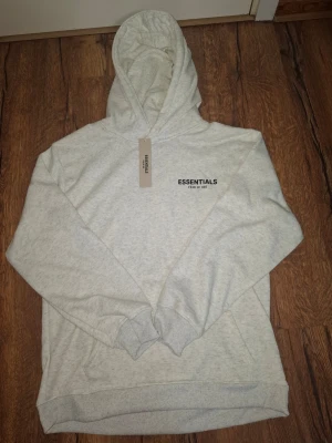 Grå Essentials Fear of God hoodie - Ljusgrå hoodie från Essentials Fear of God med svart logga tryckt på bröstet. Klassisk modell med huva och ribbade muddar vid ärmslut och nederkant. Perfekt för en avslappnad och trendig streetwear-look.