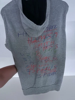 Grå hoodie med färgglad text - Säljer en grå hoodie med huva och färgglada texttryck i rött, blått och grönt på ryggen. Hoodien har långa ärmar, ribbade muddar och en avslappnad passform. Perfekt för dig som gillar streetwear och unika detaljer. Använt typ 5 gånger.