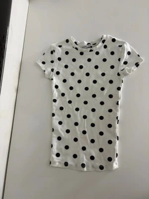 Vit prickig t-shirt från Bershka - Vit t-shirt från Bershka med svarta prickar. Klassisk rund halsringning och korta ärmar. Tillverkad i mjuk bomull, perfekt för en avslappnad och trendig look.