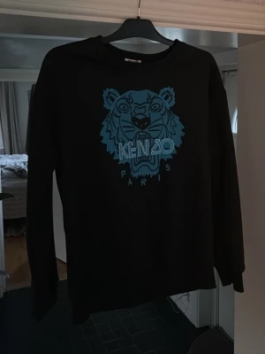 Kenzo tröja NY - Svart sweatshirt från Kenzo med ikoniskt blått tigerhuvud broderat på bröstet och texten 'KENZO PARIS'. Tröjan har rund halsringning och långa ärmar. Perfekt för dig som gillar streetwear och vill sticka ut med en klassisk design. Aldrig använd 