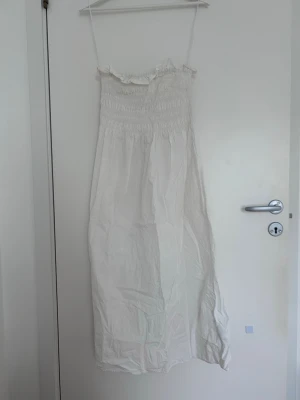 Vit långklänning med smock - Luftig vit långklänning med smala axelband och smock upptill. Klänningen har en rak passform och volangdetalj vid överdelen. Perfekt för varma dagar och enkel att styla med accessoarer.