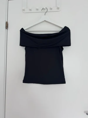 Svart offshoulder topp - Stilren svart offshoulder topp med bred krage som vilar över axlarna. Toppen har en tight passform och är perfekt för dig som vill ha en enkel men snygg look. Tillverkad i ett mjukt och stretchigt material.