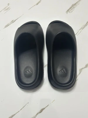 Svarta Yeezy slides från Adidas - Säljer ett par svarta Yeezy slides från Adidas med klassisk öppen design och bred rem över foten. Tillverkade i ett mjukt och flexibelt syntetmaterial för extra komfort. Perfekta att glida i hemma eller till och från träningen.