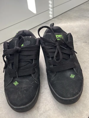 Svarta DC sneakers med gröna detaljer - Storlek 40 men passar om du har 38/39 också! Svarta sneakers från DC med breda svarta skosnören och gröna DC-loggor på tungan, sidan och framtill. Skorna har en låg profil och är tillverkade i mockaliknande material med textilfoder. Snygg streetstil med gröna grafiska detaljer på sidan.