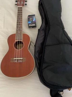 Ukulele med väska och stämapparat - Säljer en brun ukulele i trä med fyra strängar, svart gigbag och en Joyo JMT-03 clip-on stämapparat/metronom med färgdisplay. Perfekt för dig som vill börja spela eller ta med instrumentet på språng.