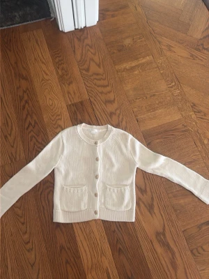 Krämvit kofta från Gina Tricot - Söt krämvit kofta från Gina Tricot med rund halsringning, ribbade muddar och två fickor framtill. Koftan har dekorativa knappar i guldton och är stickad i mjukt material. Perfekt att slänga över en topp för en chill look. Storlek 146/158 