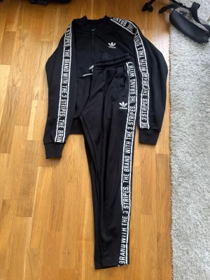 Svart Adidas tracksuit med text - Säljer ett svart tracksuit från Adidas med vita detaljer. Både jacka och byxor har texten 'THE BRAND WITH THE 3 STRIPES' längs ärmar och ben samt Adidas-logga på bröst och lår. Jackan har dragkedja och byxorna har dragsko i midjan. Tillverkad i mjukt, sportigt material.
