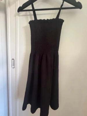 Svart sommarklänning från RYVLS - Säljer en svart kort klänning från RYVLS med smock upptill och tunna axelband. Klänningen har en enkel och stilren design som passar perfekt till varma dagar. Tillverkad i ett mjukt och lätt material som känns skönt mot huden.