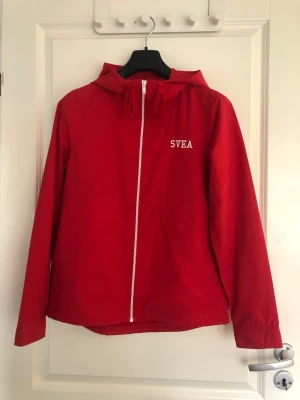 Röd vindjacka med huva från SVEA - Snygg röd vindjacka från SVEA med vit dragkedja och logga på bröstet. Är i storlek 158/164 men passar mig som har storlek XS/S