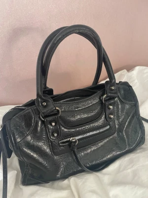 Svart handväska med dragkedjedetaljer - Snygg svart handväska i glansigt konstläder med dubbla handtag och silverfärgade dragkedjor. Väskan har en cool, lite rockig look med dekorativa nitar och en framficka med dragkedja. Perfekt för dig som gillar en edgy stil och vill ha plats för allt du behöver. Ovh det får plats med ganska mycket men är inte för stor och inte för liten. Pris kan diskuteras 💕
