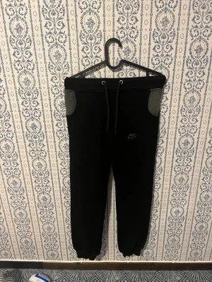 Svarta mjukisbyxor från Nike - Svarta mjukisbyxor från Nike med grå detaljer vid fickorna och dragsko i midjan. Byxorna har resår vid bensluten och en liten Nike-logga på vänster ben. Perfekta för chill dagar eller träning.